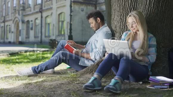年轻人拿着书坐在树下看着手提电脑的女孩优柔寡断视频的预览图