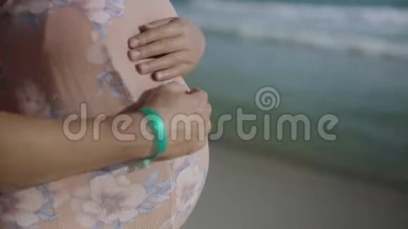 年轻的孕妇站在蓝色海洋附近的海滩上呼吸靠近手和腹部暑假假期视频的预览图