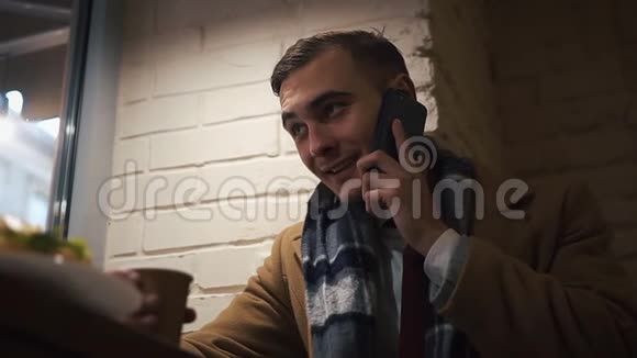 一个年轻人坐在靠窗的咖啡馆里从纸杯里喝咖啡然后打电话打电话那家伙穿着衣服视频的预览图