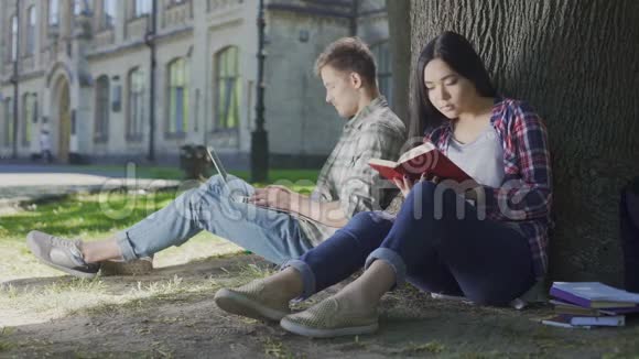 一个手提电脑的男人坐在树下看着女孩看书视频的预览图
