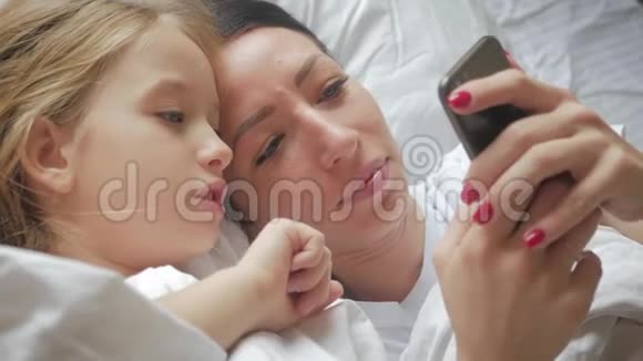 年轻漂亮的妈妈和她可爱的女儿在家里躺在床上用智能手机自拍微笑着视频的预览图