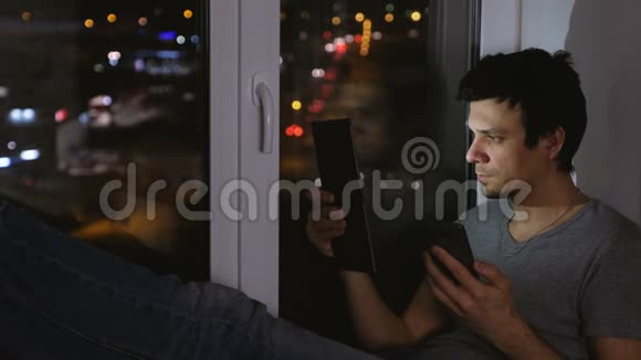 男人在看平板电脑和手机在黑暗中坐在窗台上视频的预览图