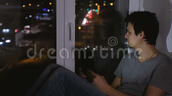 在黑暗的夜晚男人坐在窗台上看着街道使用平板电脑上网视频的预览图