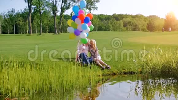 一对美丽的情侣拿着五颜六色的气球坐在河岸上的慢动作视频视频的预览图