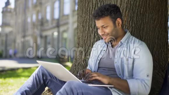 跨国青年坐在树下在笔记本电脑上打字微笑着聊天视频的预览图
