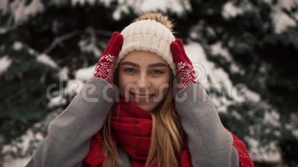 穿着暖和衣服的年轻漂亮女孩站在圣诞树旁戴着帽子看着镜头视频的预览图
