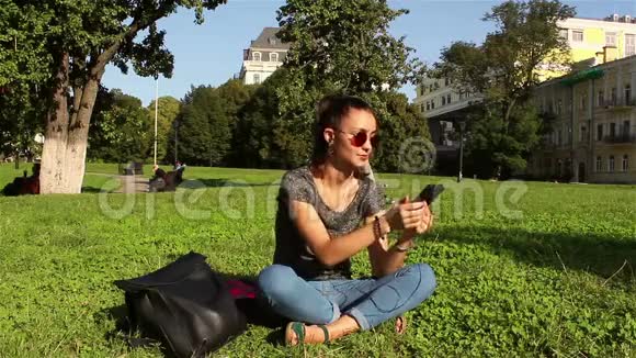 美丽的女孩坐在公园的草地上通过手机里的耳机听她最喜欢的旋律视频的预览图