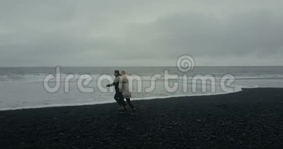 科普特乘坐冰岛毛衣在这对年轻夫妇附近飞行男人和女人在黑色的火山海滩上奔跑视频的预览图