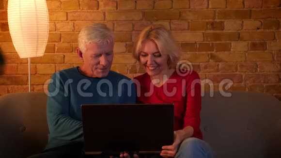 在舒适的公寓里坐在沙发上的高级幸福夫妇在笔记本电脑上浏览的特写镜头视频的预览图