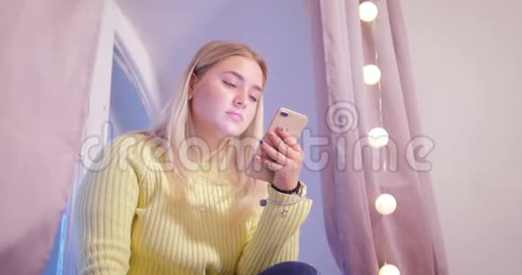 一个穿着黄色毛衣的金发女孩正在房间里坐着电话背景是粉红色的窗帘和视频的预览图