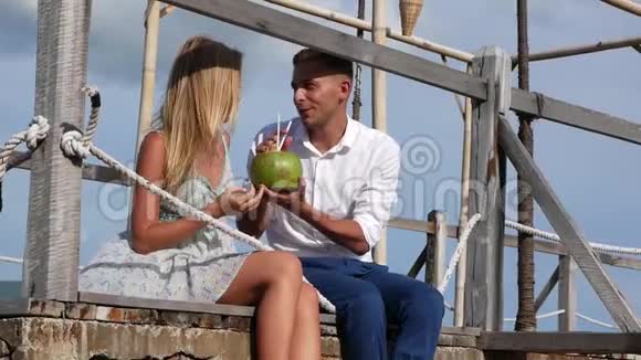 年轻的幸福夫妇坐在木墩上笑着喝着蓝天背景上的椰子浪漫浪漫视频的预览图