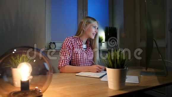 微笑的女人晚上在电脑上工作穿着格子衬衫坐在小灯木桌旁点燃的微笑的女性视频的预览图