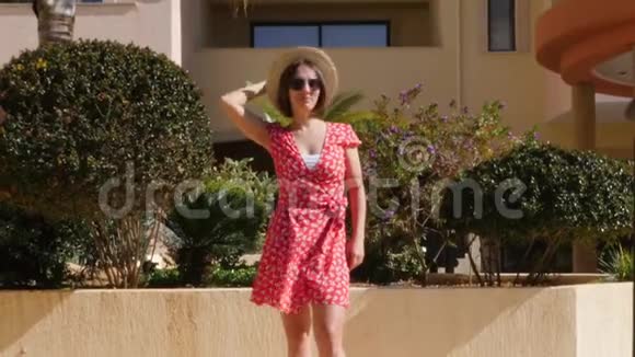 美丽的白种人女人戴着太阳镜戴着帽子穿着红色的裙子带着棕榈树走向花园里的摄像机视频的预览图