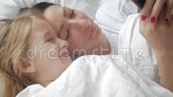 年轻漂亮的妈妈和她可爱的女儿在家里躺在床上用智能手机自拍微笑着视频的预览图