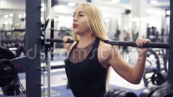 强壮的年轻金发女人穿着黑色衣服漂亮的运动身材在健身房用杠铃做蹲式锻炼视频的预览图