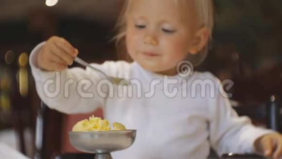 婴儿在餐馆吃食物坐在高椅上的金发碧眼的孩子从盘子里拿出一茶匙冰淇淋然后拿走视频的预览图