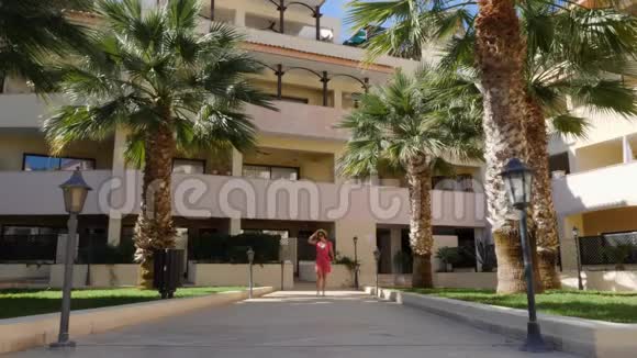 迷人的美女走在酒店花园的镜头前穿着红色连衣裙戴着帽子戴着太阳镜摆着姿势棕榈树视频的预览图