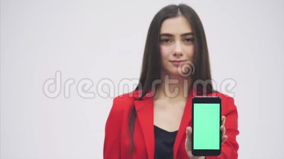 年轻漂亮的女孩穿着一件红色夹克停留在灰色背景上在此期间手持黑色手机绿色视频的预览图