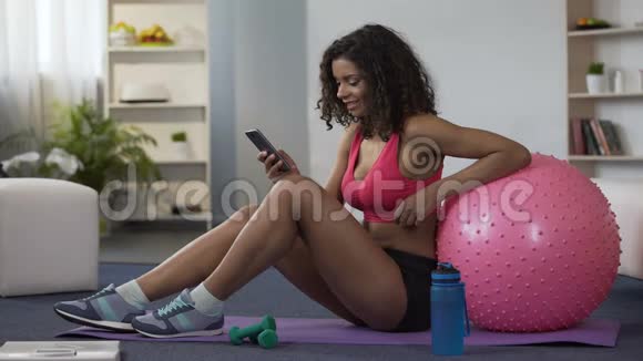 穿着健身服的双种族妇女坐在地板上使用手机申请视频的预览图