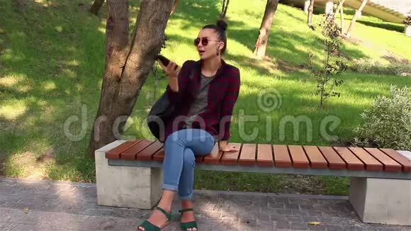 年轻漂亮的女孩在智能手机里听音乐坐在公园长凳上唱歌跳舞视频的预览图