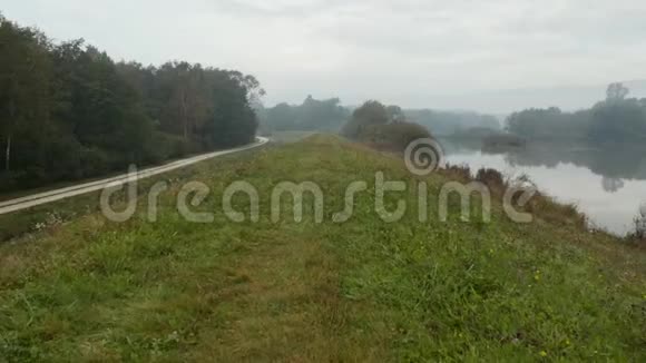 在一个雾蒙蒙的秋晨无人机沿着一座长满青草的小山和附近的反光湖射击视频的预览图