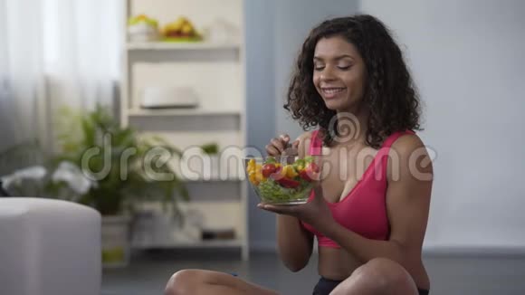 年轻健康的女人吃蔬菜沙拉坐在地板上穿着健身房的衣服节食视频的预览图