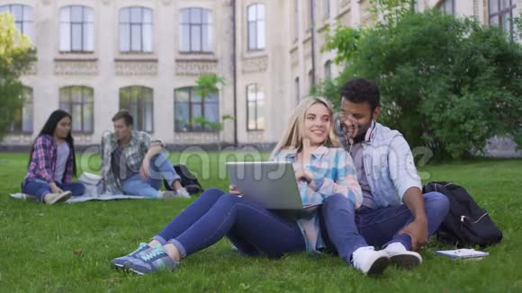 多民族学生坐在草地上用笔记本电脑观看有趣的视频视频的预览图
