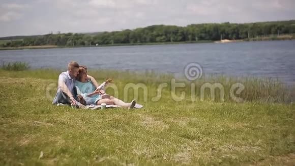 年轻的一对恋爱中的情侣坐在河岸边的绿草上视频的预览图