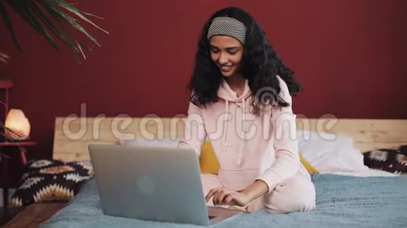 漂亮的女孩穿着粉红色睡衣家里坐在床上用笔记本电脑年轻女子微笑着聊天视频的预览图