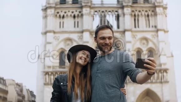 幸福的年轻夫妇站在巴黎圣母院附近法国和在智能手机上自拍照片视频的预览图