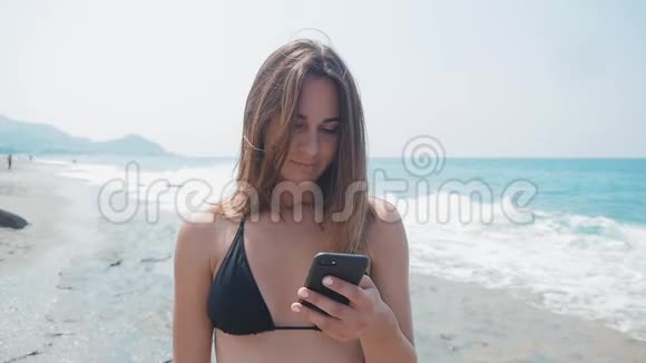 年轻的女人在海滩上使用智能手机在海上度假她穿着比基尼在放松的时候视频的预览图
