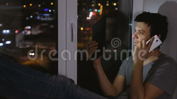 快乐的人在摆弄手机和看平板电脑在黑暗的夜晚坐在窗台上视频的预览图