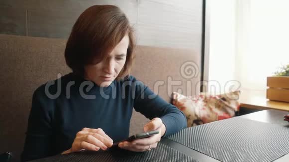女人在咖啡馆里用智能手机打短信咖啡馆里桌子旁的黑发女人坐着电话视频的预览图
