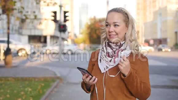 幸福的金发年轻女子正在通过耳机听音乐穿过秋天的城市瞬间视频的预览图
