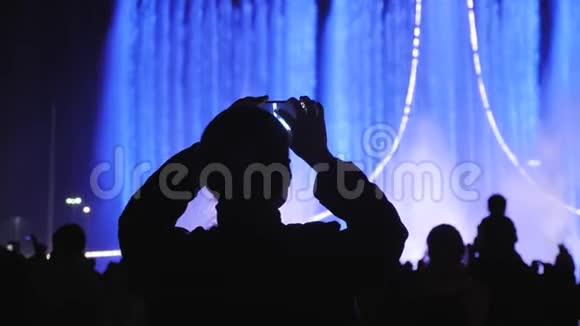 那些正在观看和拍摄明亮彩色喷泉的人的剪影人们拍一张光的照片视频的预览图