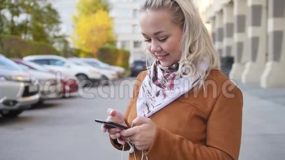 幸福的金发年轻女子正在通过耳机听音乐穿过秋天的城市瞬间视频的预览图