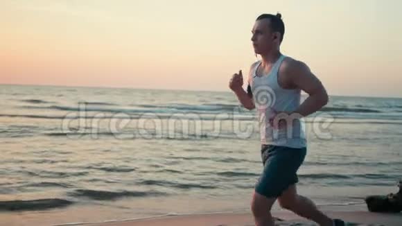 运动型男人拿着电话在沙滩上慢跑日落时分在海边跑步视频的预览图