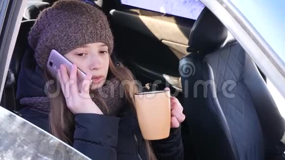 年轻漂亮的女孩坐在车前座位上打电话喝咖啡微笑视频的预览图