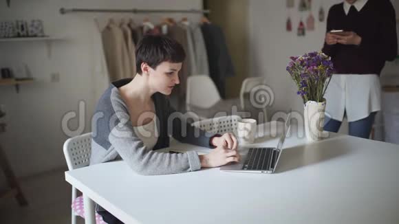 年轻女子坐在桌子旁用笔记本电脑打字女人用智能手机视频的预览图