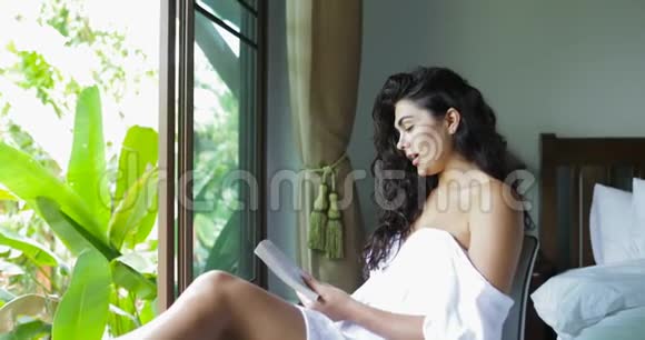 年轻女性阅读书籍快乐欢笑美丽的女孩在卧室里翻过大窗户带着热带花园的景色视频的预览图