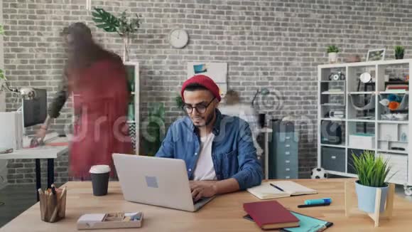 年轻男子在办公室使用笔记本电脑工作而同事在移动视频的预览图
