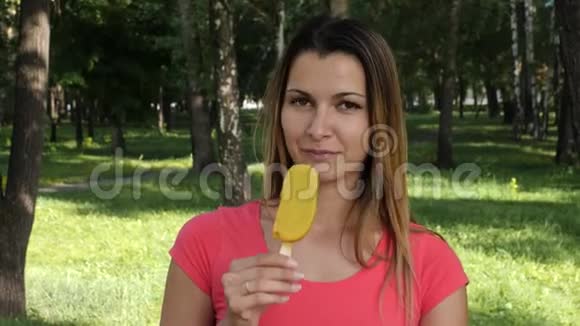 年轻迷人的女人在公园里吃冰淇淋女孩在绿树丛中吃鲜艳的冰淇淋视频的预览图