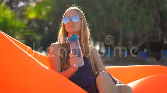 热带海滩上的年轻女子坐在充气沙发上从一个多用途的塑料瓶里喝水饮料视频的预览图