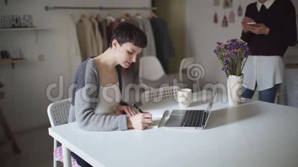 年轻女子坐在桌子旁在笔记本上做笔记女人用手提电脑视频的预览图