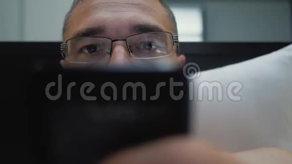 一个戴着眼镜躺在床上的男人在智能手机上交流视频的预览图