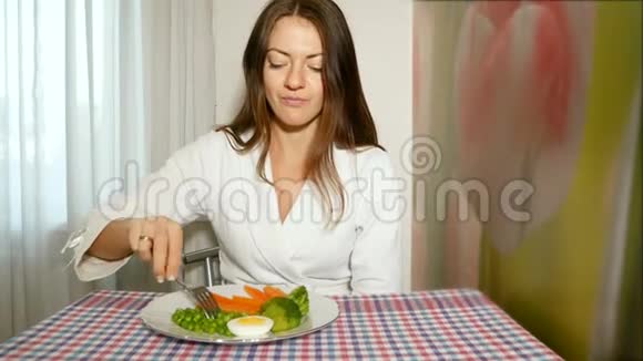 一个穿着白色长袍的漂亮女孩坐在厨房的桌子旁试着用叉子把豌豆弄绿视频的预览图
