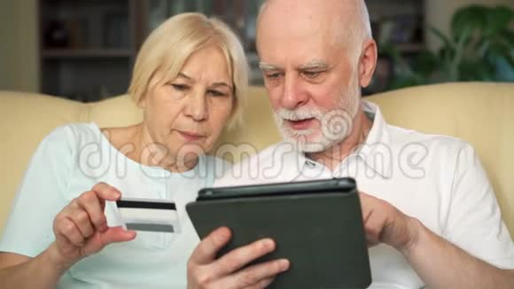 开朗好看的老两口坐在沙发上使用信用卡在线购买平板电脑视频的预览图