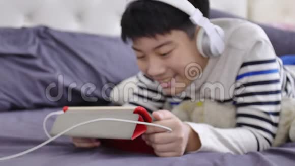 亚洲可爱的男孩在平板电脑上观看戴着白色耳机脸上带着微笑视频的预览图