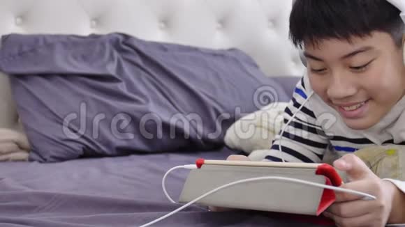 亚洲可爱的男孩在平板电脑上观看戴着白色耳机脸上带着微笑视频的预览图