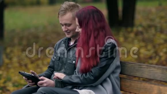 可爱的夫妇坐在秋季公园的长凳上一起使用数字平板电脑有着红色头发的年轻女子视频的预览图